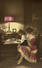 1910, Mother with child practising writing, child and mother at a desk in dim lamp light,