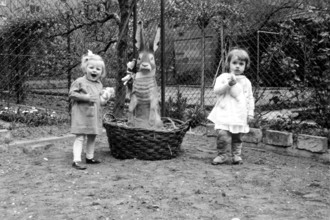 Easter bunny basket girl, 1920, Two small children standing in a garden next to a large basket with
