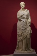 Full-length portrait of an ancient female statue in elaborate drapery against a red background,