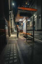 Person waiting at an illuminated bus stop in a quiet city street at night, London, United Kingdom,