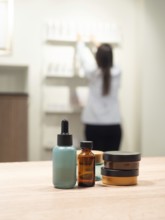Skincare and cosmetic product bottles are on a table with a blurred scientist arranging items in