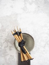 Multiple bamboo paint brushes tied with a black ribbon are resting on a ceramic bowl on a textured