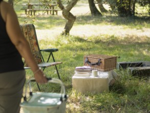 Woman bringing picnic basket to an outdoor setup with a wicker basket and camping chair