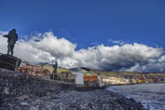 Coastline with statues and houses under a cloudy sky along a rocky shore, Candelaria Tenerife