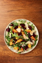 A bowl of salad contains beetroot, orange slices, pine nuts, and leaves. It is placed on a wooden