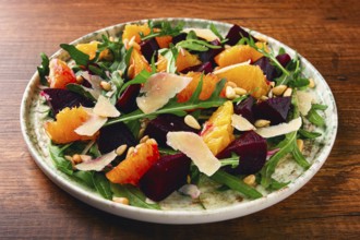 A bowl of salad contains beetroot, orange slices, pine nuts, and leaves. It is placed on a wooden