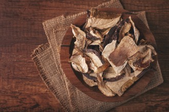 A collection of dried mushrooms fills a wooden bowl placed on a cloth surface. The mushrooms are