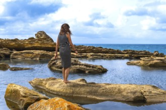 Woman standing barefoot on textured sandstone by tidal pools, facing the vast sea under a cloudy