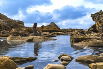 Woman sitting alone on a rock surrounded by shallow calm water and eroded rock formations,