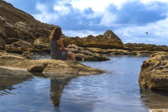 Woman sitting on ancient rocks by a tranquil tidal pool, watching the calm sea and rugged coast of