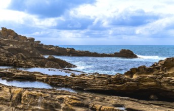 Ocean waves crashing on a rugged rocky coastline with tide pools reflecting the cloudy sky,