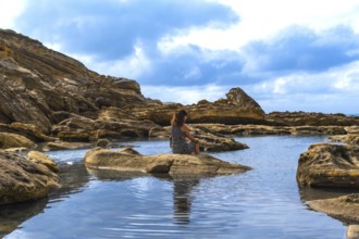 Woman sitting on a rock in a calm tide pool. Reflecting on the water. Surrounded by rugged coastal