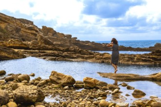 Woman standing barefoot on a rock, observing the peaceful blue sea framed by natural rocky outcrops