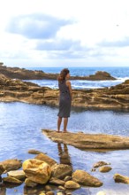 Woman standing barefoot on a rock formation, observing the ocean and the rugged coastline with