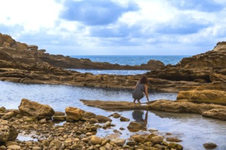 Woman crouching by a rocky tide pool on a rugged coastline, exploring the shallow water with the