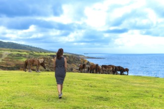 Woman walking along the verdant grass of jaizkibel mountain, observing wild horses grazing freely
