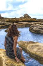 Woman sitting on coastal rock formations, enjoying the calm water and rugged landscape of