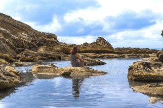 Woman contemplating the serene environment, sitting on a rock by a tranquil ocean rock pool with a