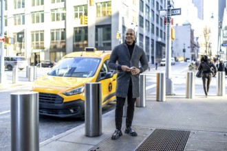 Young businessman stands on a manhattan sidewalk holding a smartphone beside a yellow taxi, smart