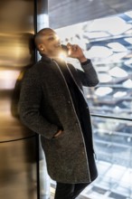 Man in an elegant coat making a phone call, leaning against a metallic wall inside a sunlit