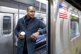 Man standing at a new york subway entrance with a takeaway coffee, looking away while waiting to