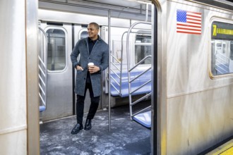 Young businessman standing on a subway car with a coffee cup, looking out through the open door,