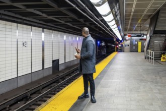 Businessman in a winter coat stands on the yellow safety strip of a subway platform, holding a