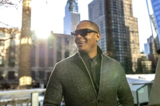 Young man smiling and looking away while wearing dark sunglasses and a gray jacket against a sunny