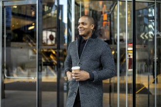 Confident young businessman standing outside a modern office building, holding a disposable coffee