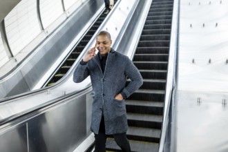 Young man standing on an escalator, holding a cellphone to his ear while smiling, representing