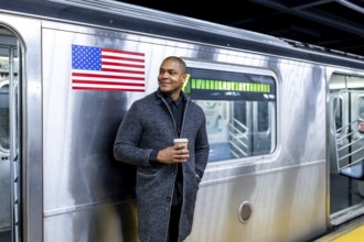 Smiling man standing on a subway platform in manhattan, enjoying a coffee while waiting for public