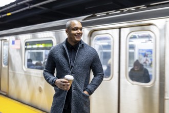 Young businessman standing on a manhattan subway platform with a take out coffee, looking away