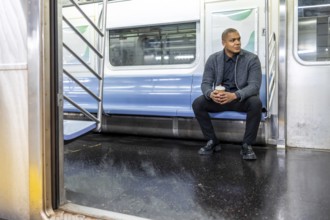Young man sitting in an empty subway car holding a coffee cup, looking thoughtfully out the window