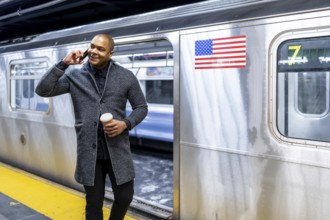 Black businessman standing on a subway platform, talking on a smartphone and holding coffee,