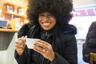 Young black woman joyfully holding a cup of coffee while smiling widely in a cozy cafe, portraying