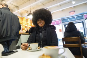 Young black woman with an afro hairstyle smiling while holding a cup of coffee at a table in a