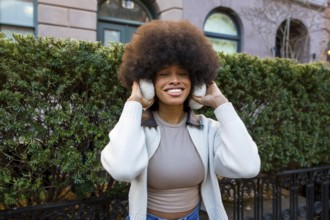 Smiling black woman with large afro hair wearing earmuffs and a sweater, standing outdoors on a