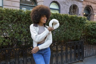 Young black woman with a large afro hairstyle smiling outdoors, holding white earmuffs while