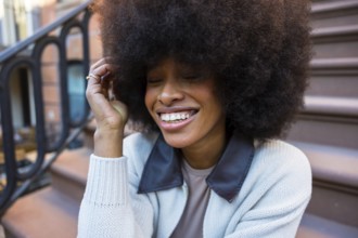 Young black woman laughing with eyes closed and hand in hair, sitting on a brownstone stoop,