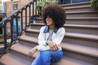 Young black woman with afro sitting on harlem brownstone stoop, smiling and relaxed, enjoying sunny