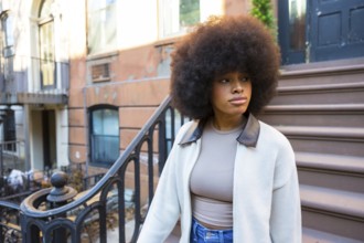 Young black woman with natural afro hair standing on an urban brownstone stoop, looking away while