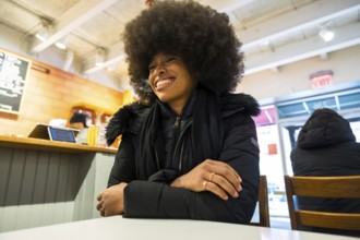 Smiling black woman in a winter jacket chatting happily inside a bustling manhattan coffee shop,