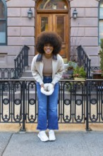 Smiling woman with afro hair holding a white furry hand warmer, standing on a sidewalk in front of