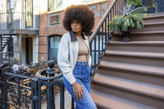 Stylish young woman with afro hair posing confidently on the brownstone steps of an urban building,