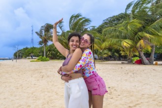 Two happy young women friends embracing on a tropical sandy beach with palm trees and a clear blue