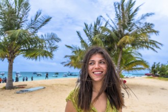 Young woman smiling and looking away, standing on a sandy beach with palm trees and a turquoise