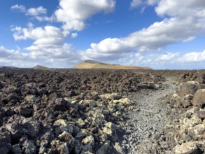 Hiking trail through lava rock to Caldera Blanca volcano against blue sky with white clouds, Mancha