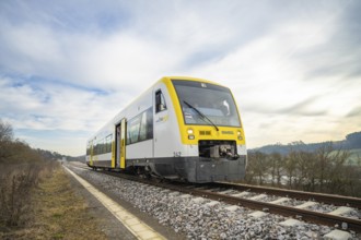A yellow and white train travels on rails through an open landscape under a cloudy sky, taking off