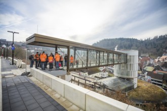 People in orange vests stand on a glass bridge with a view of a city and hills, taking off the
