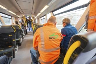 Men talking on a train, one wearing an eye-catching orange AVG jacket, acceptance trip of the